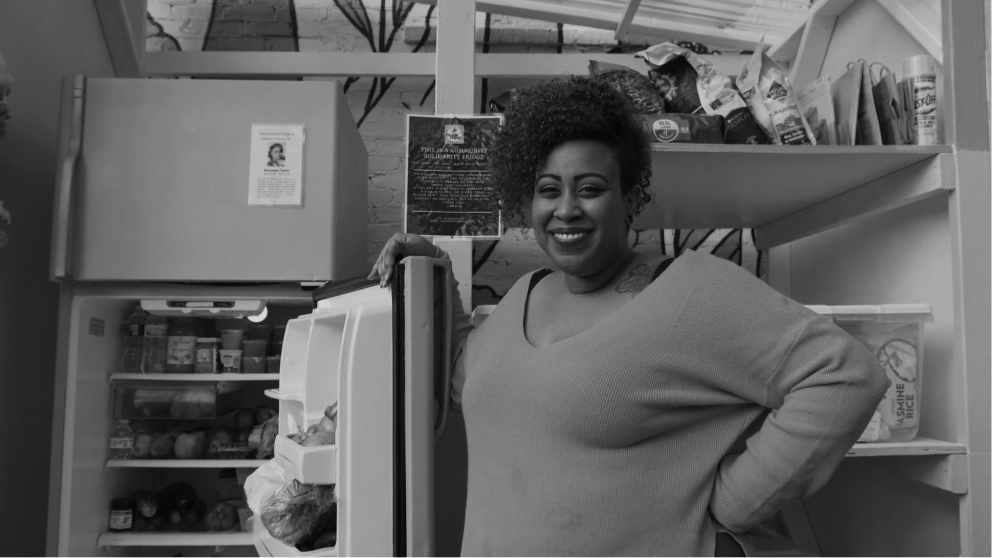 A woman standing beside a stocked community refrigerator in a black-and-white campaign image