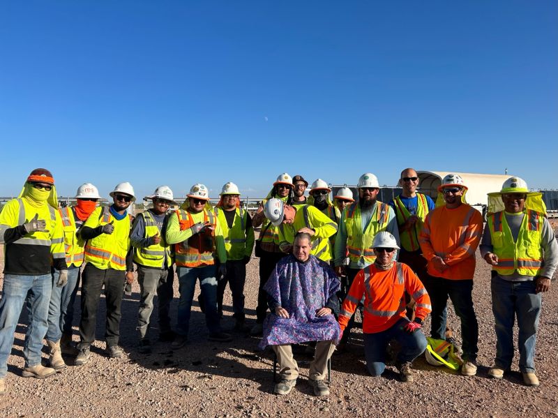 Matt Campbell seated in a haircut cape, surrounded by the solar project crew