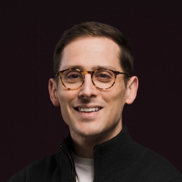 Matt Epstein wearing round glasses and a dark zip-neck sweater against a dark background