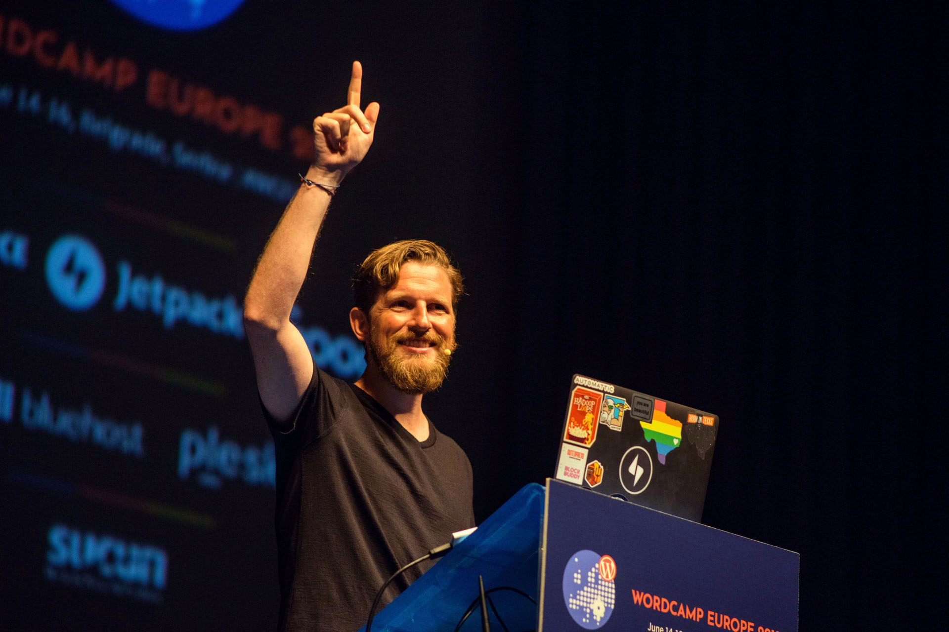 Matt Mullenweg raising a finger while speaking behind a laptop at WordCamp Europe 2018
