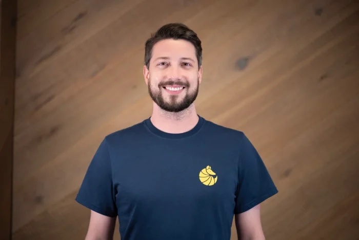 Maximilian Birner smiling in a navy Boxhub T-shirt against a wooden wall