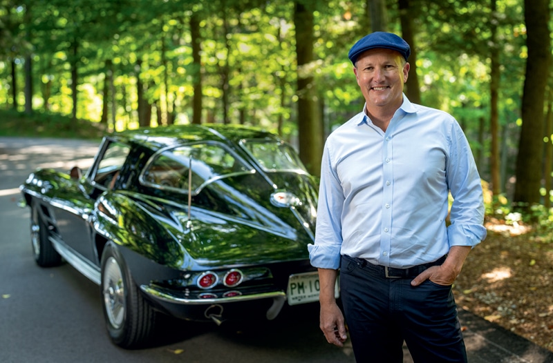 McKeel Hagerty standing beside his dark green 1963 Chevrolet Corvette on a wooded road