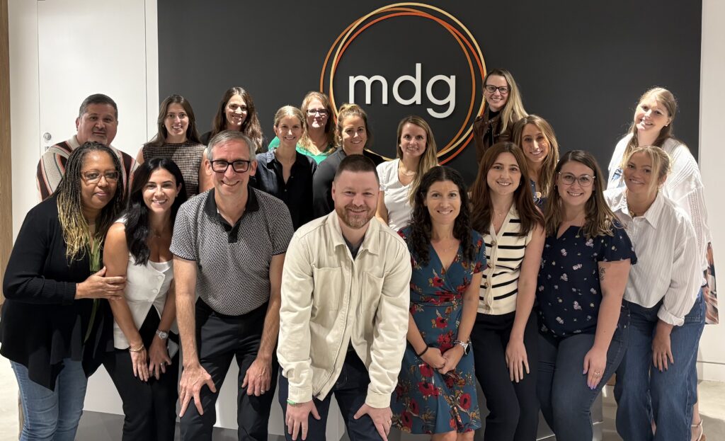 mdg team members gathered for a group photograph in front of the agency’s wall logo