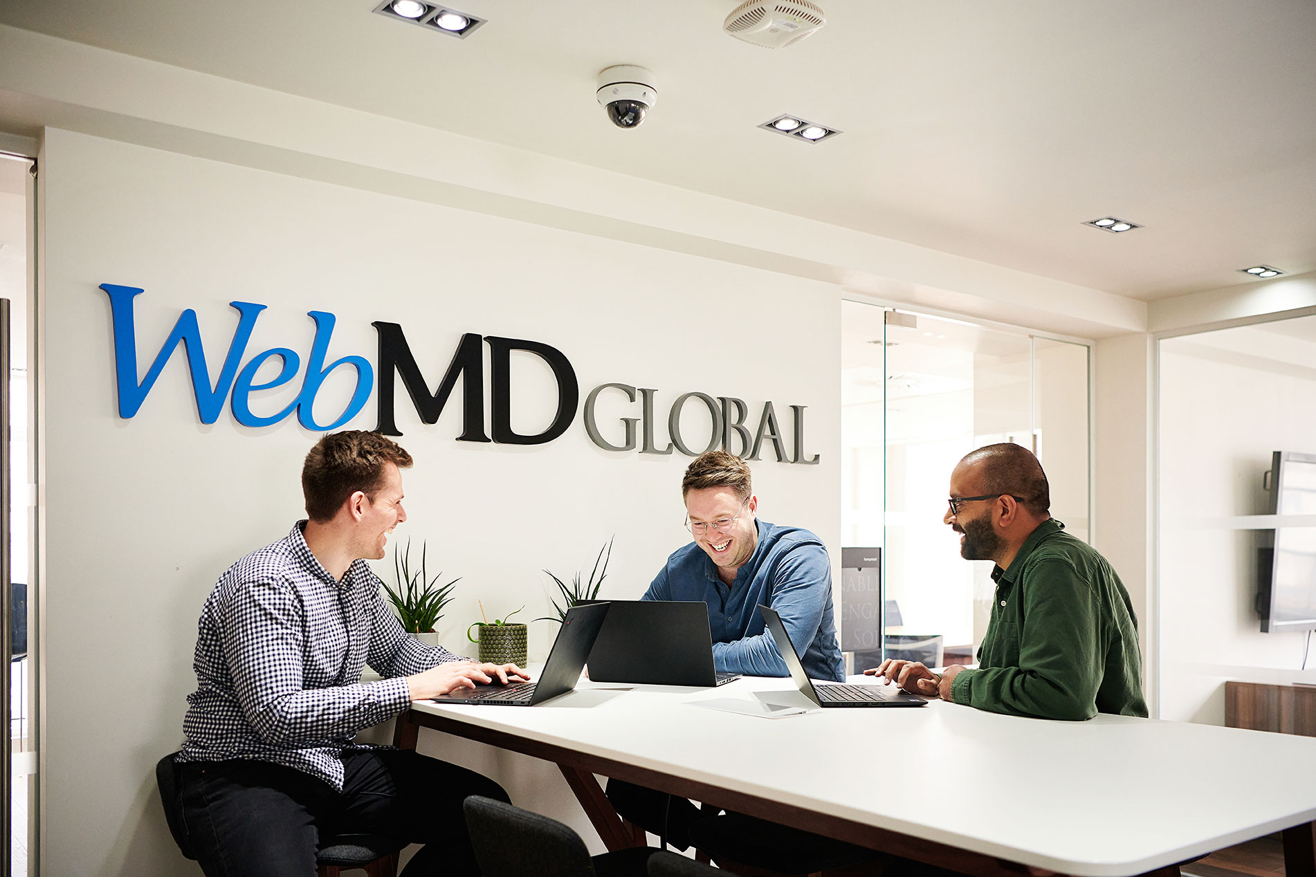 Three people with laptops seated around a table beneath a WebMD Global sign