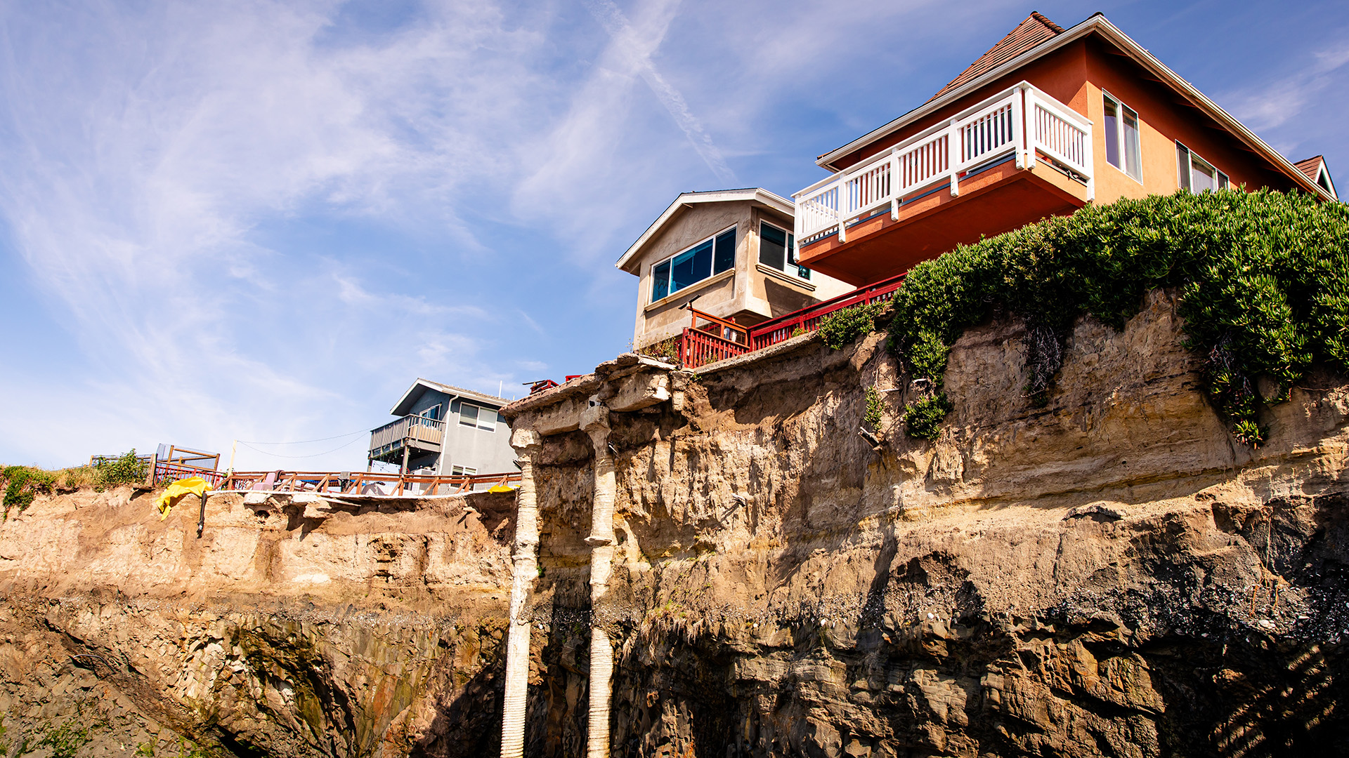 Homes standing above a visibly eroded coastal cliff under a blue sky