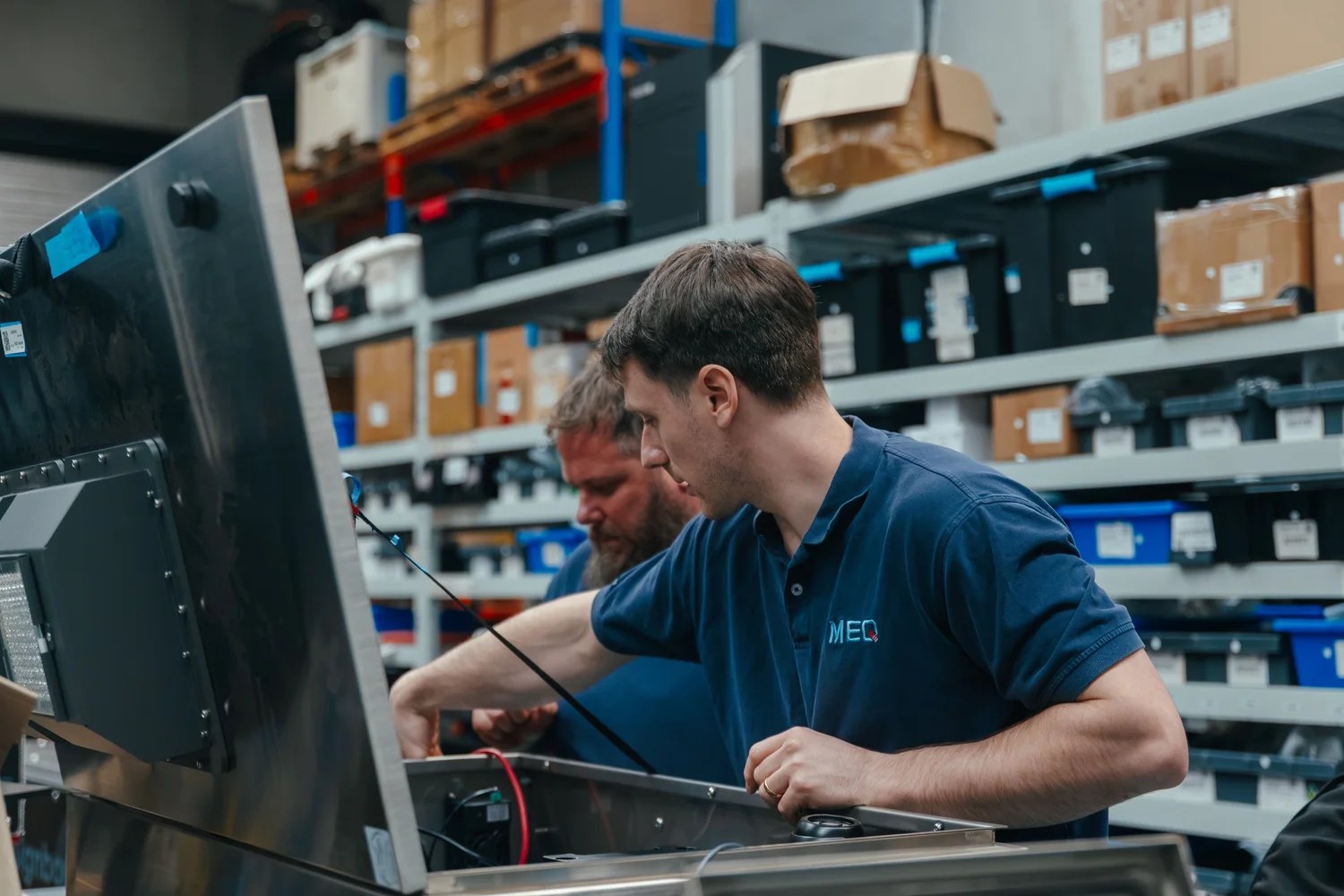 Two MEQ Solutions staff in navy MEQ polo shirts assemble a stainless-steel measurement device in a workshop lined with parts bins
