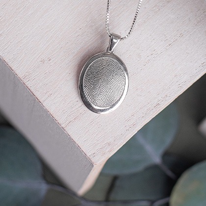 Thumbies rimmed fingerprint pendant in silver-colored metal, resting on a wooden surface