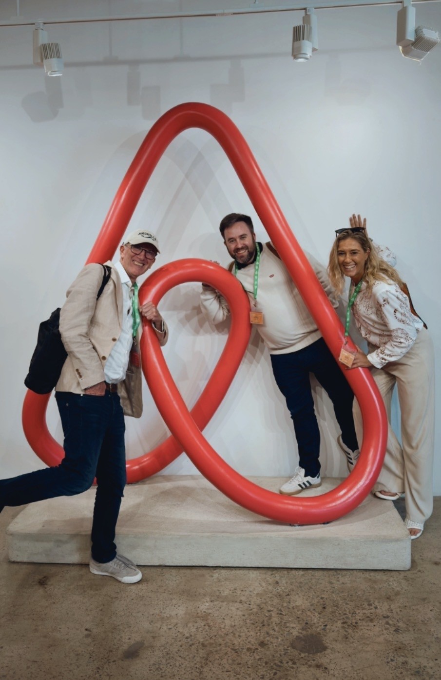Three people posing beside a large red Airbnb symbol in the photograph published with MIC Global’s partnership announcement