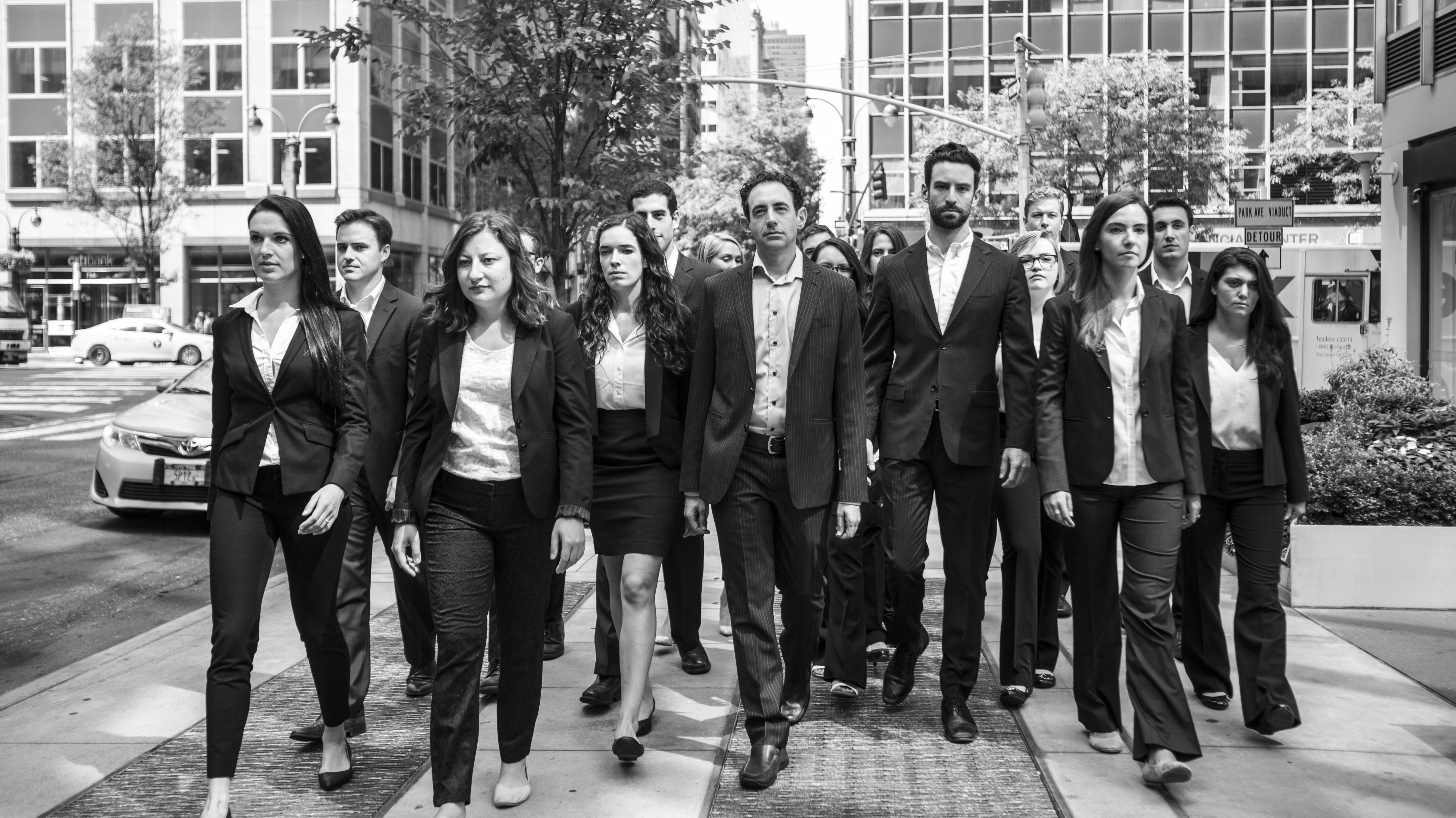 An archival black-and-white photograph of the Group Gordon team walking along a New York street, with Michael Gordon in the center.