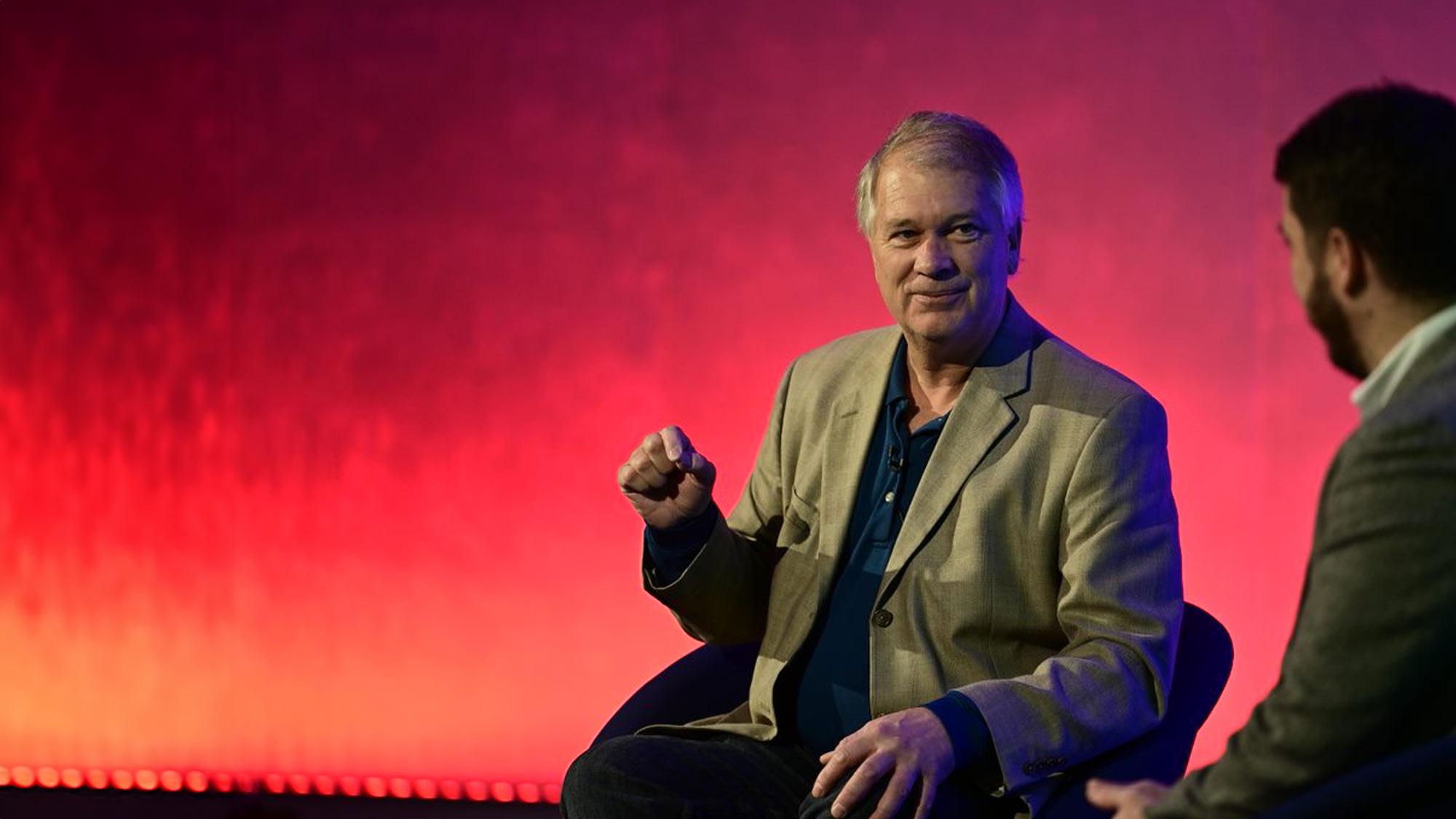 Michael Gorton in a tan jacket, seated in conversation on a stage lit red and orange