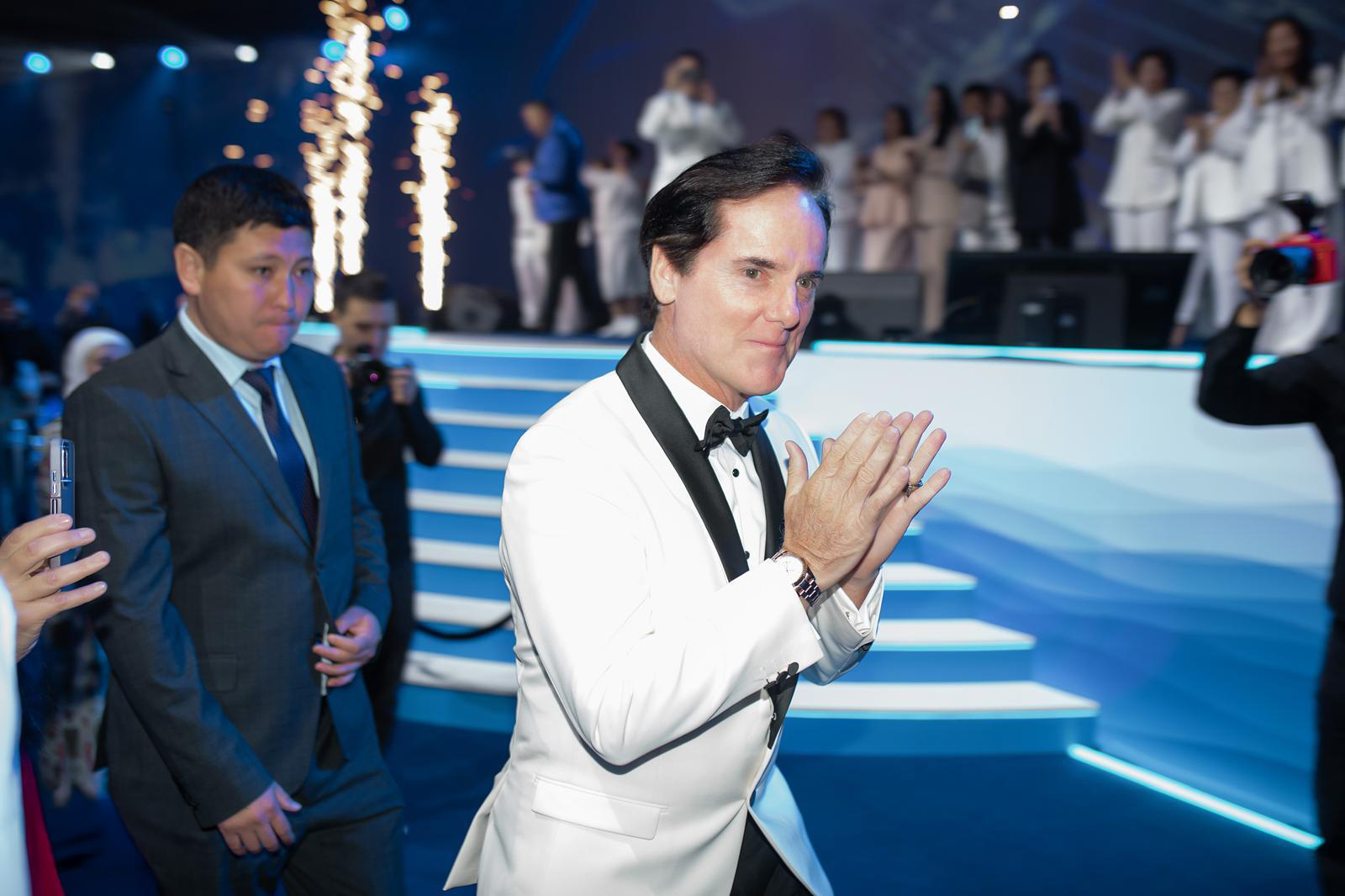 Michael Hutchison in a white dinner jacket applauding beside an event stage
