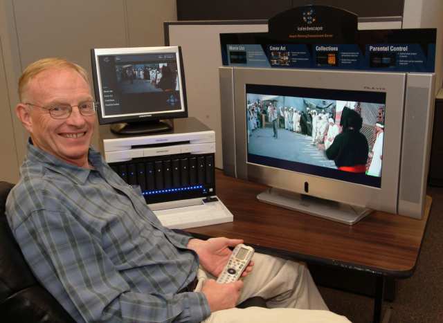 Michael Malcolm seated beside a Kaleidescape demonstration system, holding a remote control