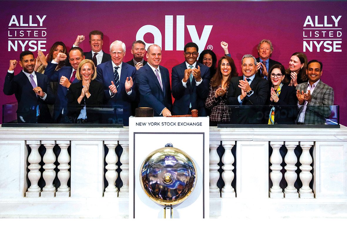 Michael Rhodes and colleagues celebrating on the New York Stock Exchange balcony beneath Ally signs