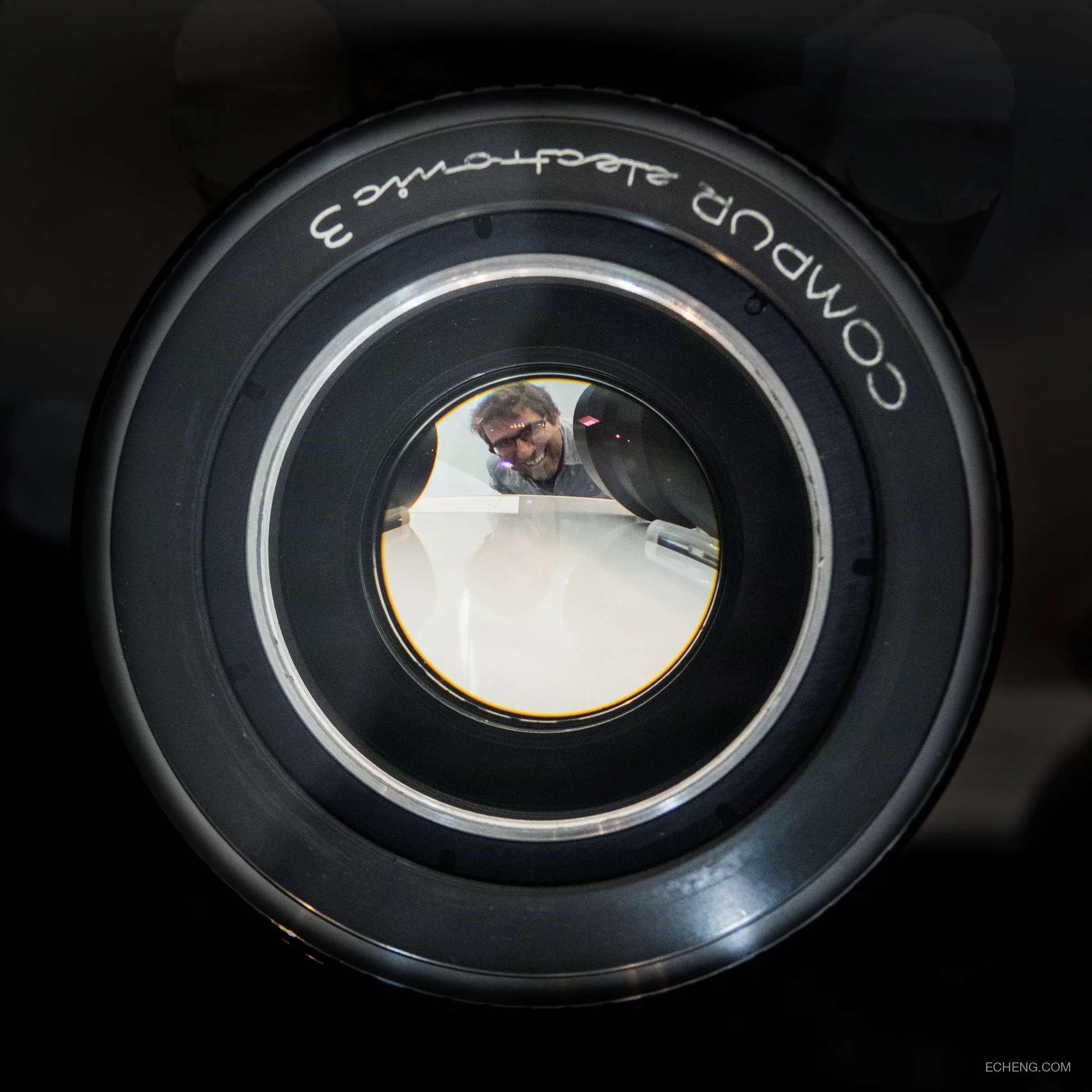 Michael Topolovac smiling through the glass of a large camera lens at a museum