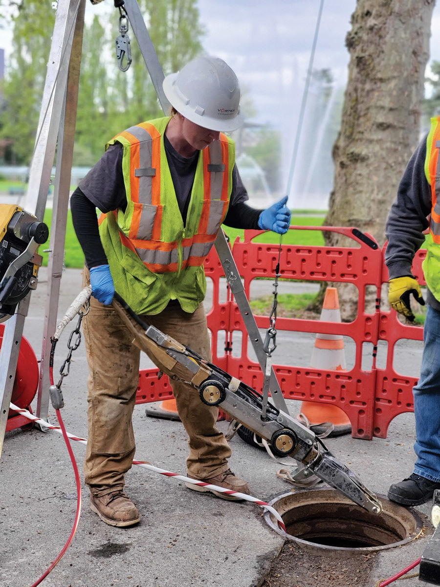 A trenchless technician lowers equipment into a manhole at an active worksite