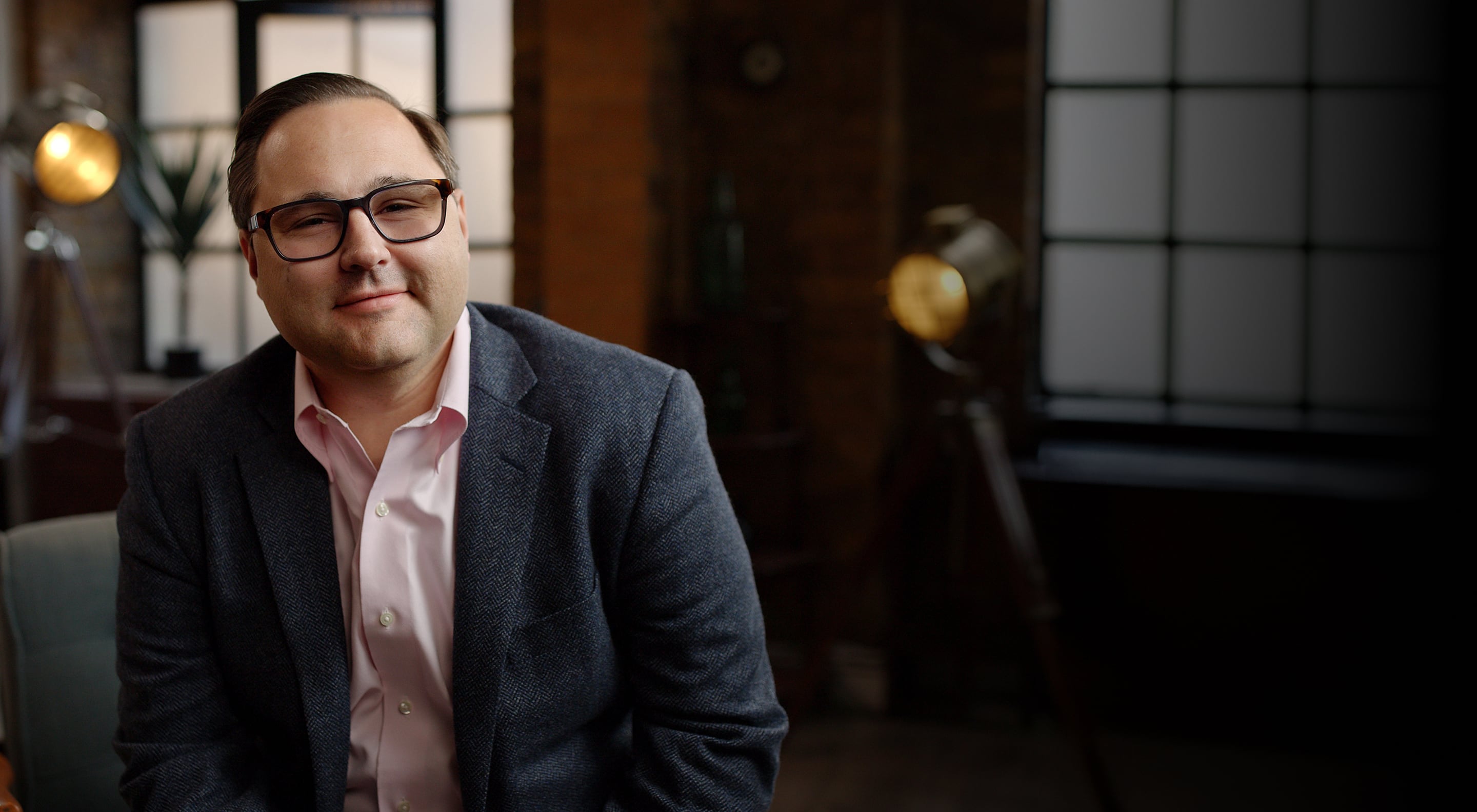 MichaelAaron Flicker seated in a jacket and glasses in an interior with studio-style lights.