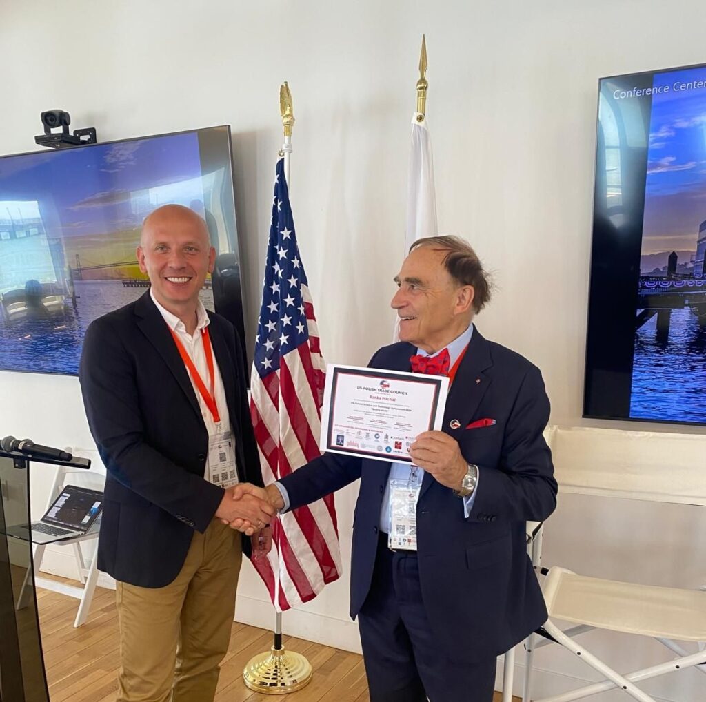 Michał Bańka, left, smiling during a handshake beside the American and Polish flags at the 2024 US-Poland symposium