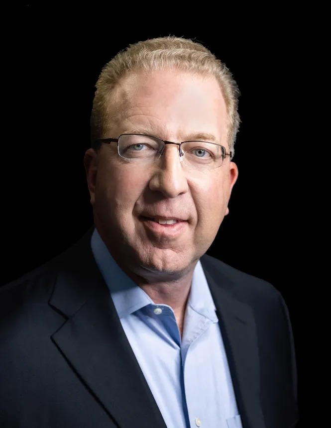 Mike Wolfel in a dark jacket and light blue shirt against a black background