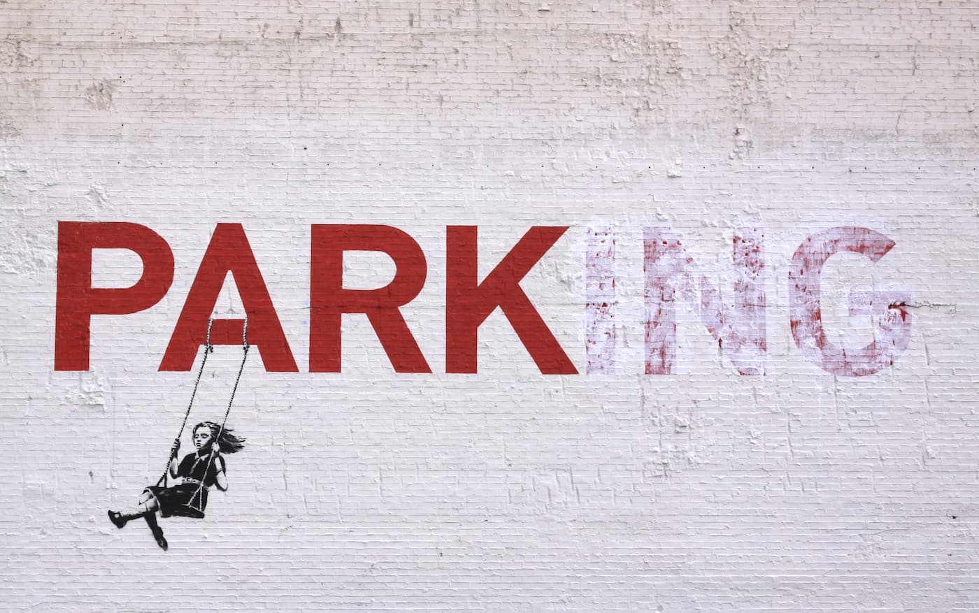 Banksy’s Girl on a Swing suspended from the A of a PARKING sign on a white brick wall