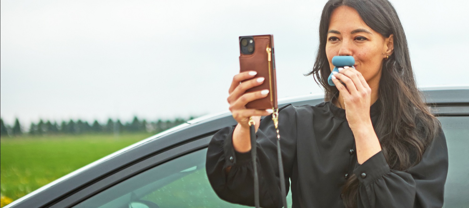 A woman uses a blue Keepr breathalyzer while holding her smartphone beside a parked car.