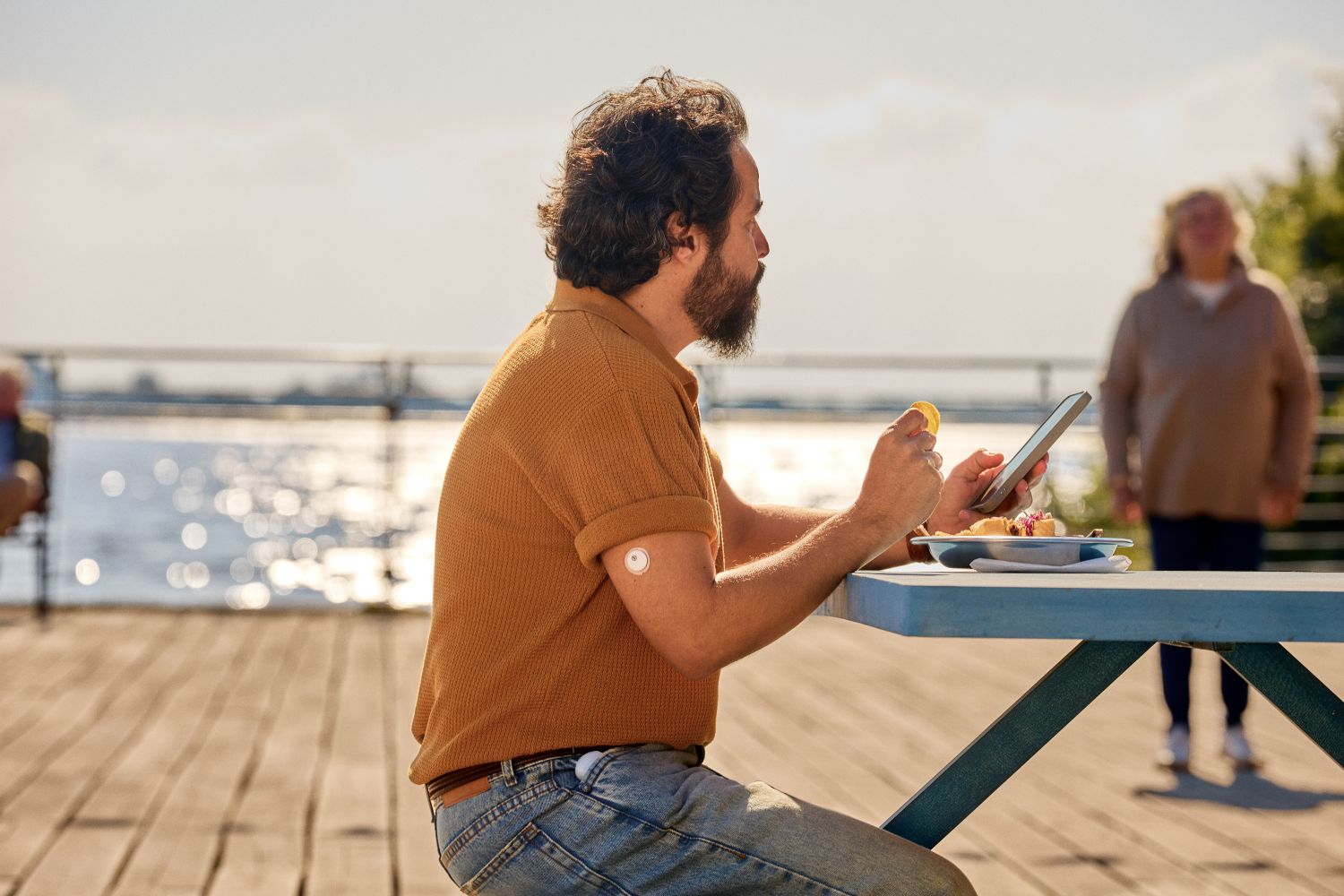 Man at an outdoor table checking his phone, with an Instinct glucose sensor on his arm and a Flex pump at his waist