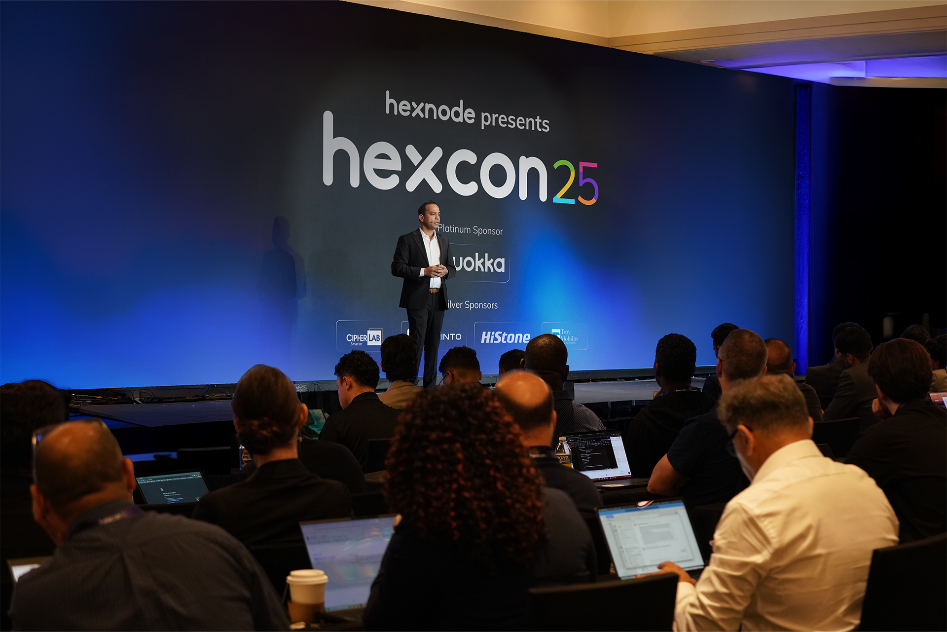 Apu Pavithran speaking on stage at HexCon25, with attendees using laptops