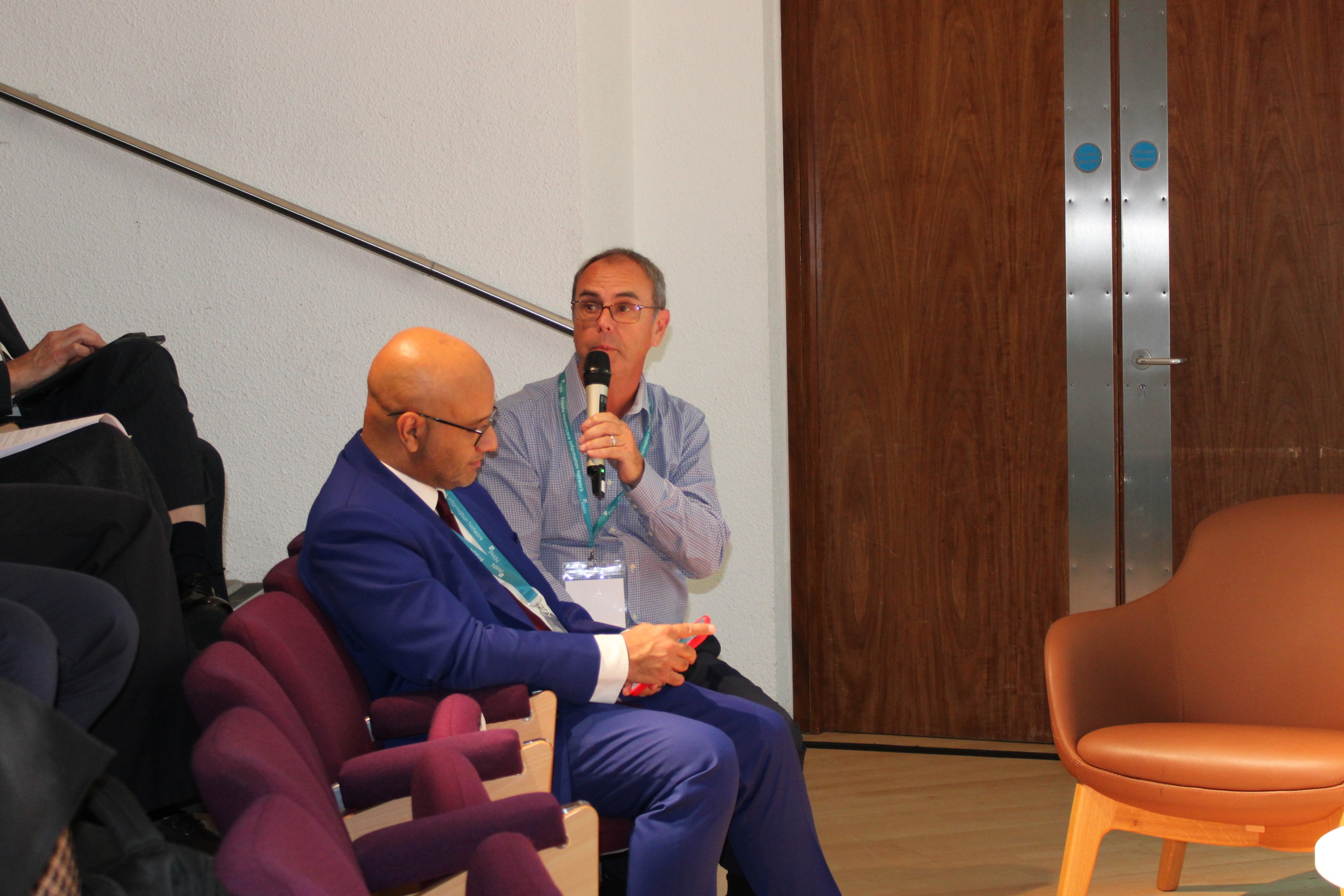 Mohammad Al-Ubaydli, seated in a blue suit, listens beside an audience member holding a microphone.
