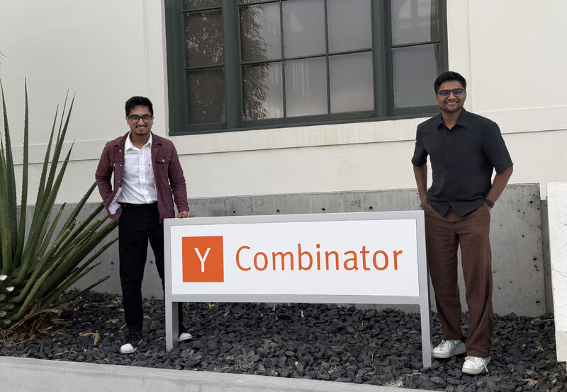Mohit Gupta and Saksham Gupta standing on either side of a Y Combinator sign