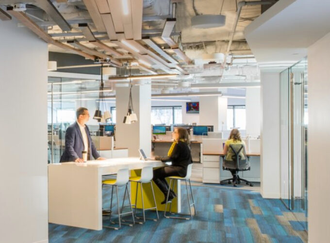 People working and talking in a bright Monex office with shared desks and yellow stools