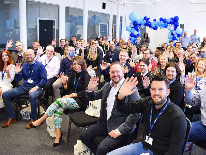 Moorepay colleagues seated together, waving toward the camera beneath blue and white balloons
