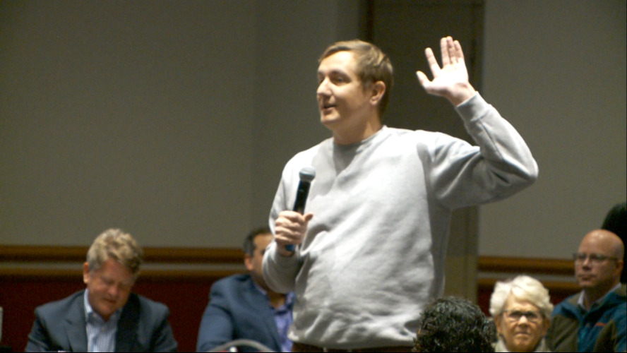 Morgan Davis stands with a microphone and raises one hand while speaking at a Utah technology policy event