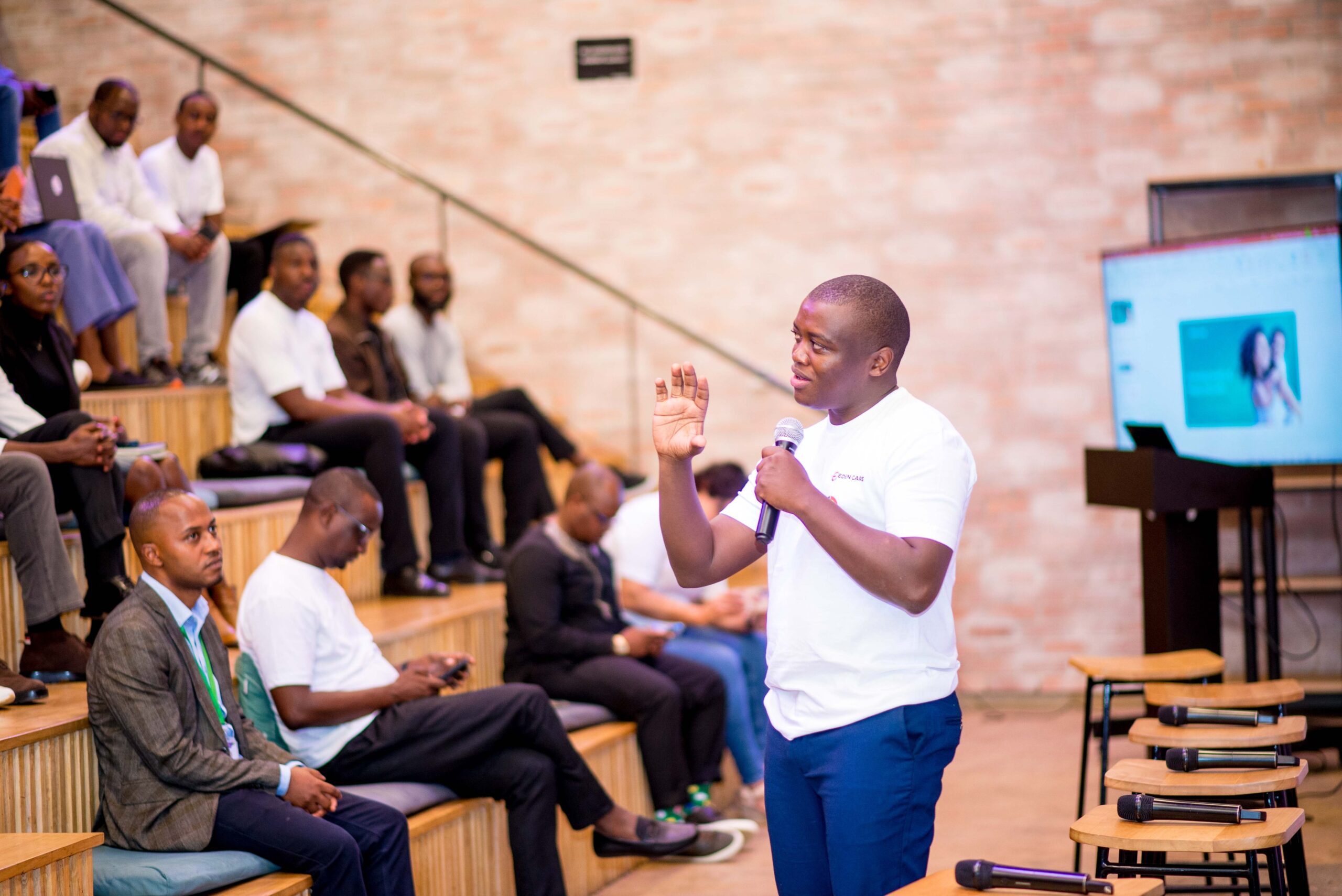 Moses Mukundi holding a microphone while addressing an audience seated on tiered benches