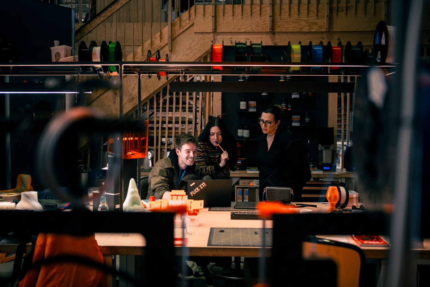 Three people working around a laptop among 3D printers and filament spools