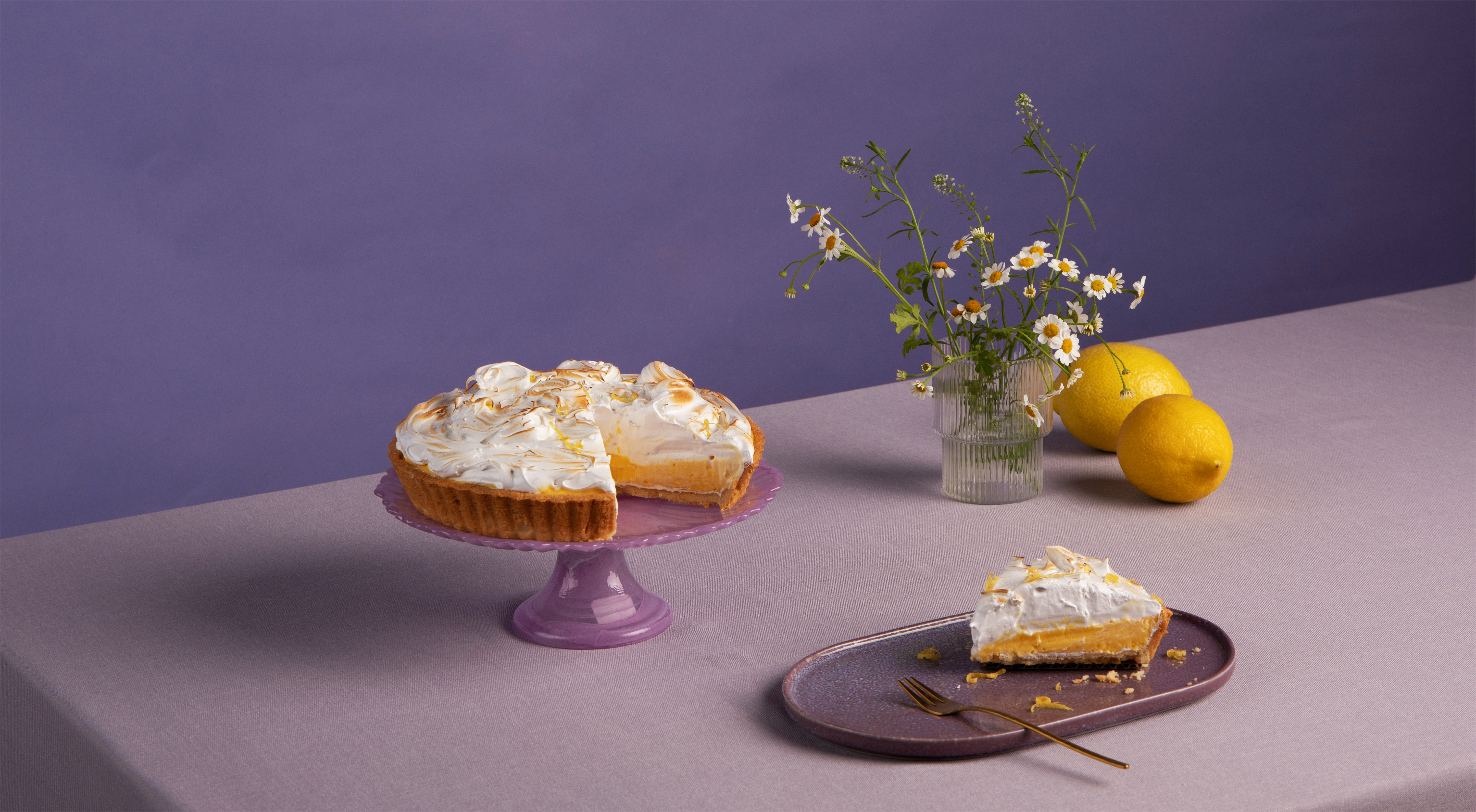 Lemon tart and a plated slice against a purple backdrop, with lemons and flowers