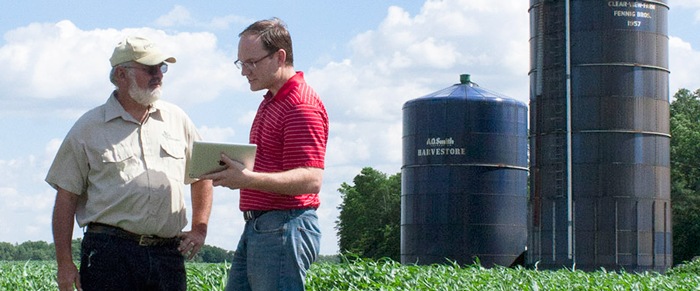 Two men talking beside a cornfield and farm silos, one holding a tablet