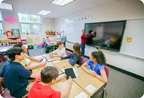 Students gathered around a table while a teacher uses a Promethean interactive display