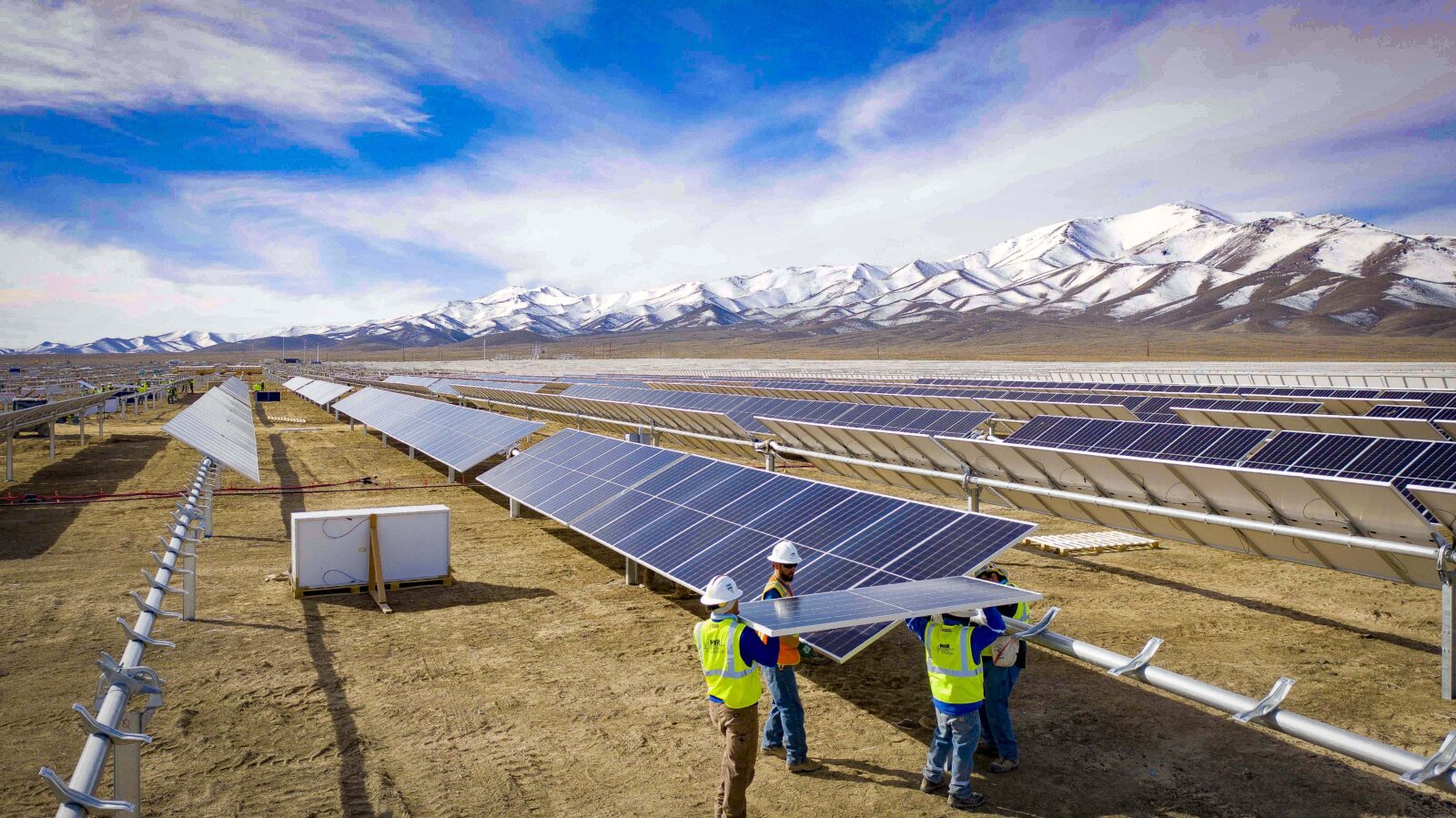 Workers carrying a solar panel between rows of panels at Battle Mountain, with snow-covered mountains behind them