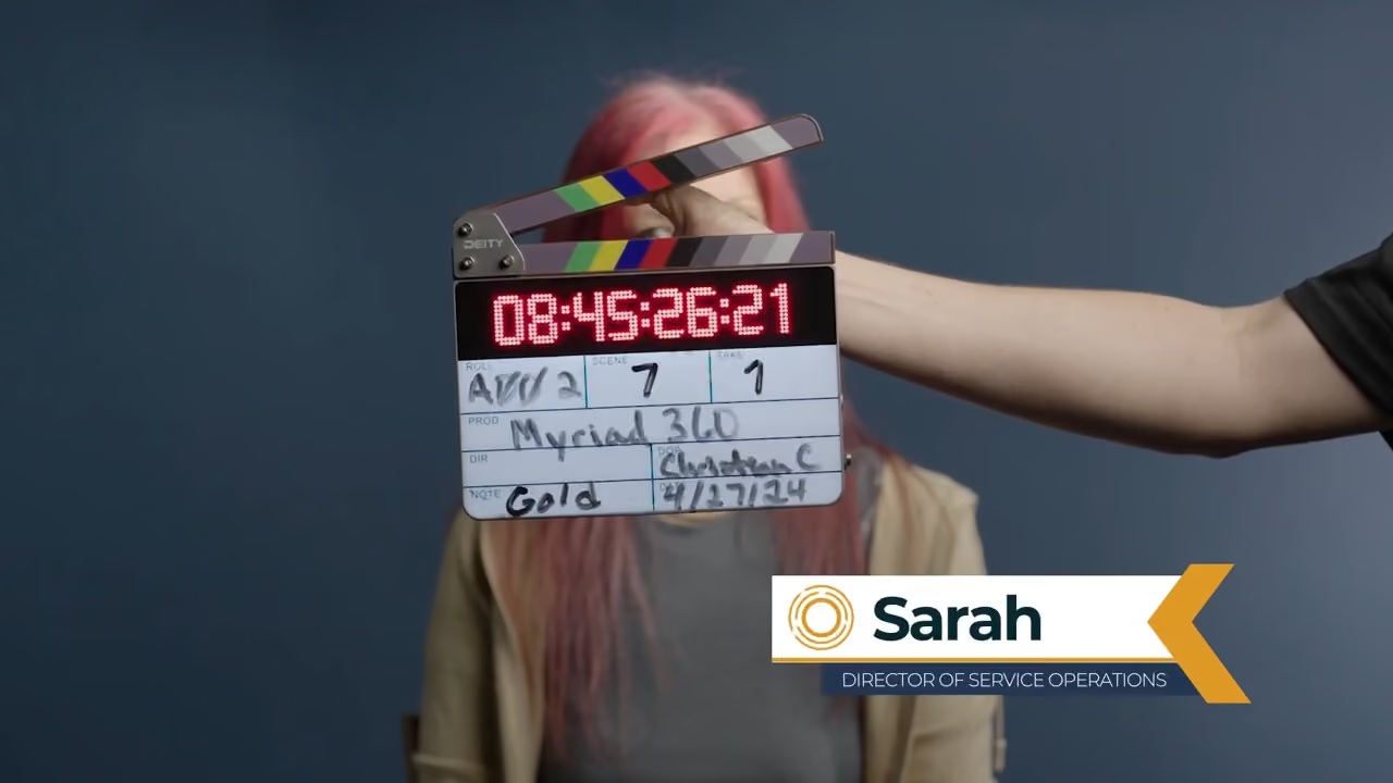 A clapperboard in front of Sarah, identified in a Myriad360 staff video as director of service operations
