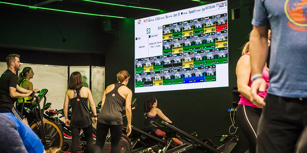 Gym members exercising below a large Myzone screen showing their live heart-rate tiles