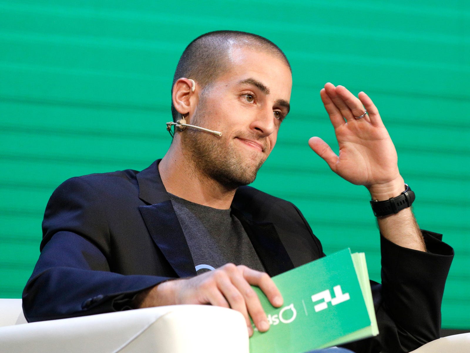 Nadav Shoval speaking onstage, seated with a headset microphone and raising one hand against a green backdrop