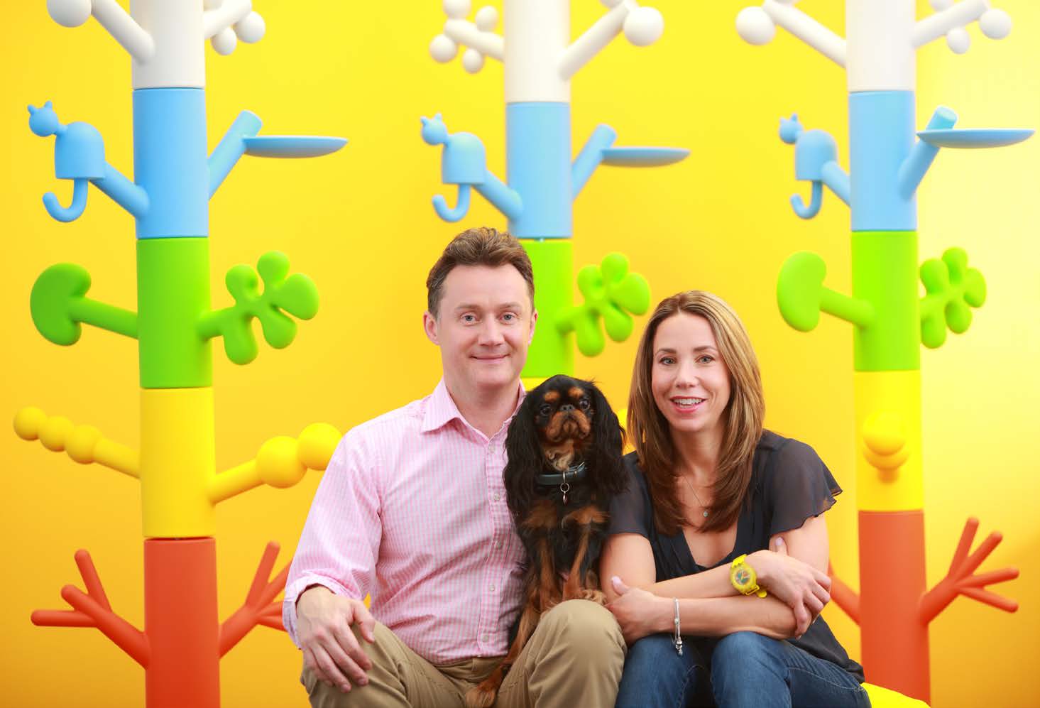 Chris Ashton and Natasha Ashton seated with their dog Monty against a yellow wall and colorful sculptures