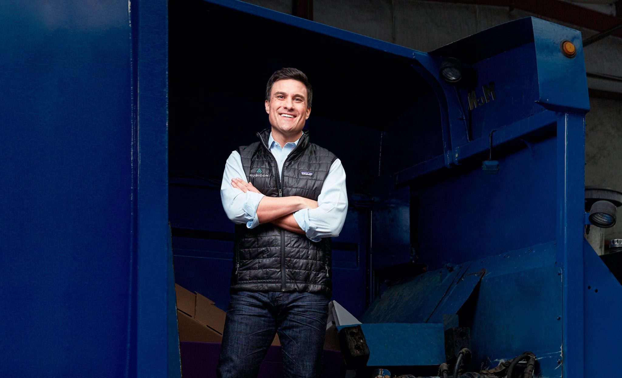 Morris in a Rubicon vest standing beside a blue industrial waste compactor