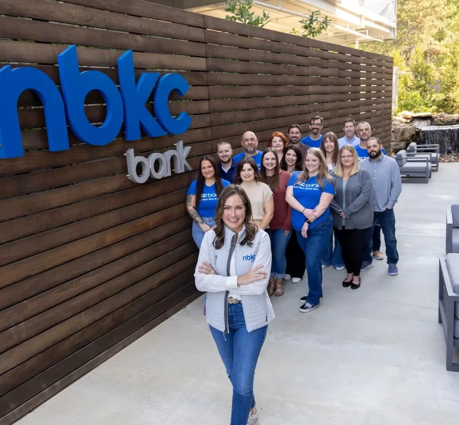 A group of nbkc bank employees gathered beside a wooden wall outside the office