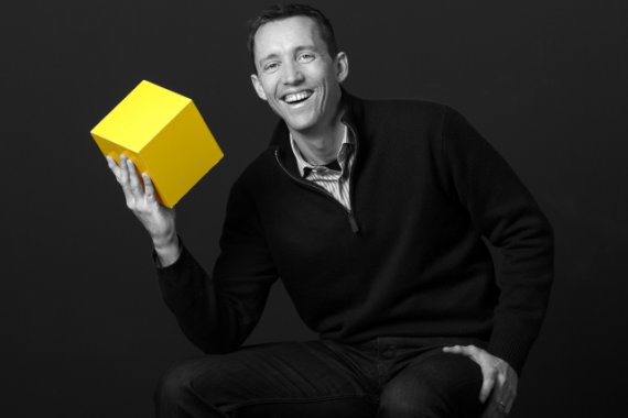 Neil Watson seated, smiling and holding a bright yellow cube