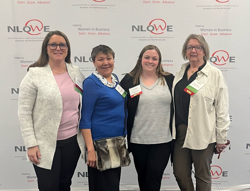 Four women wearing event badges stand together in front of a NLOWE backdrop.