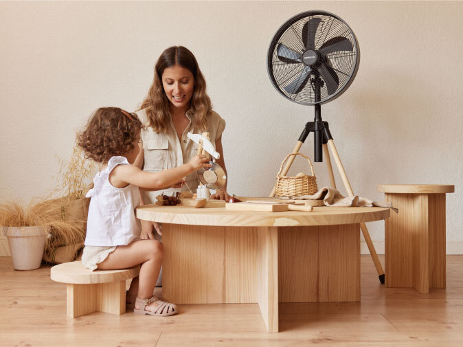 A woman and child playing at a wooden table with a Newlux standing fan behind them