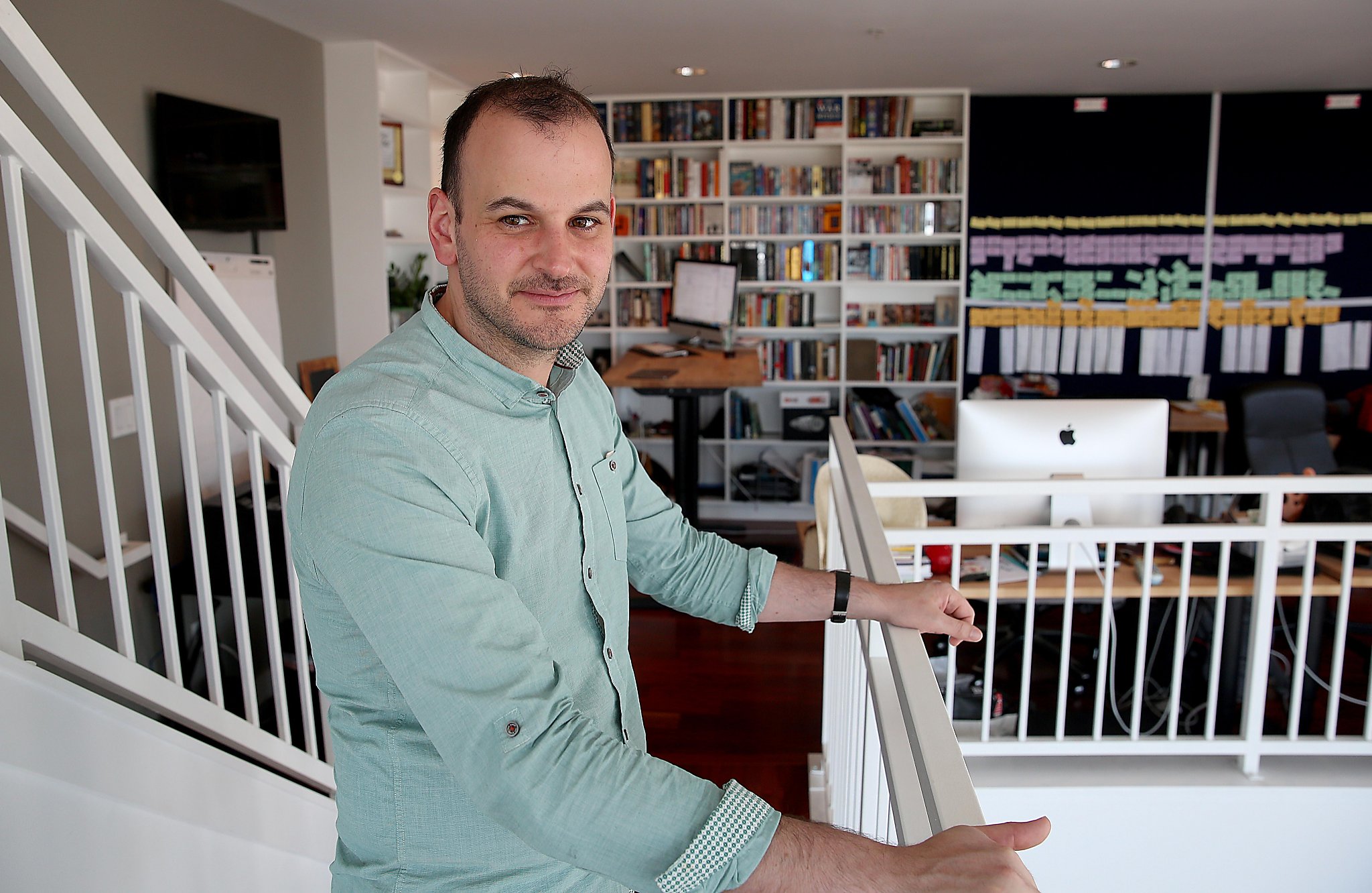 Nicholas Tommarello beside a white railing in his San Francisco home office, with bookshelves and a planning board behind him