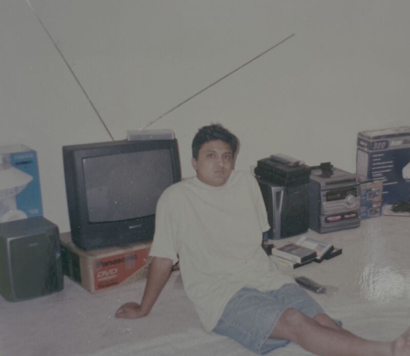 An earlier photograph of Nikhil Gupta seated on the floor beside a television and stereo