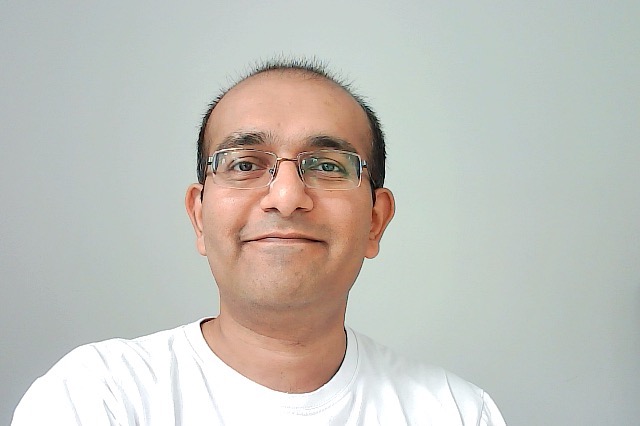 Nilay Oza wearing glasses and a white T-shirt, photographed against a pale background