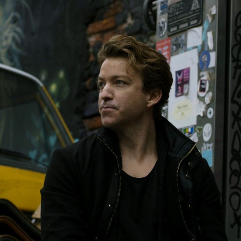 Nils Schneider outdoors, looking to his right beside a sticker-covered wall